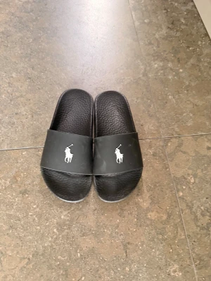 Svarta slides från Polo Ralph Lauren - Säljer ett par svarta slides från Polo Ralph Lauren med den klassiska loggan i vitt på ovandelen. Tillverkade i syntetmaterial med räfflad sula för bra grepp. Perfekta för chill dagar eller till stranden.