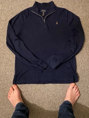 Mörkblå halvzip från Polo Ralph Lauren - Mörkblå långärmad tröja från Polo Ralph Lauren med halvzip och klassisk broderad logga på bröstet. Tröjan har ribbade muddar och en stilren, enkel design som passar till det mesta. Perfekt för lager på lager och snygg till jeans.