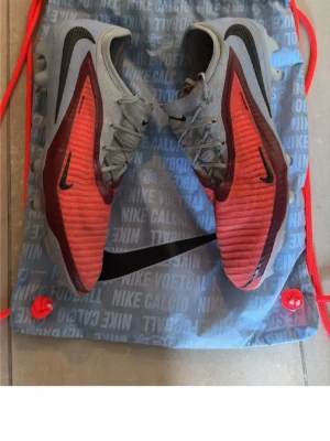 Nike blå och röda fotbollsskor - Säljer ett par Nike fotbollsskor i grått och rött med svarta detaljer och snörning. Skorna har en modern design med mönstrad ovandel och tydlig Nike-logga på sidan. Perfekta för dig som vill sticka ut på planen.