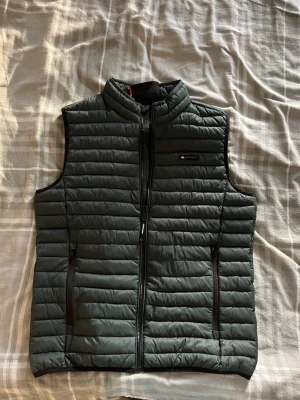 Mörkgrön dunväst från Superdry - Snygg mörkgrön dunväst från Superdry med quiltad design och hög krage. Västen har två dragkedjeförsedda fickor framtill och diskret logga på bröstet. Perfekt att slänga över en hoodie för extra värme och stil.