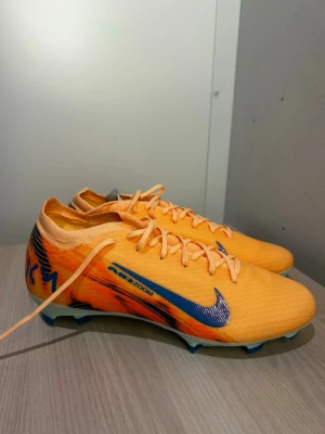 Nike orangea fotbollsskor Air Zoom - Snygga fotbollsskor från Nike i en stark orange färg med blå detaljer och logga. Skorna har snörning, låg profil och mönstrad sula för bra grepp på planen. Tillverkade i ett lätt syntetmaterial som ger bra bollkontroll och komfort. Perfekta för dig som vill sticka ut på planen.