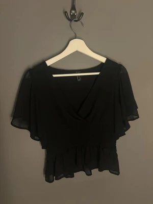 Svart omlottblus med volang - Snygg svart blus med omlott och v-ringning. Blusen har korta, vida ärmar i transparent tyg och smockad midja med volang nertill. Perfekt för dig som gillar en feminin och trendig stil.