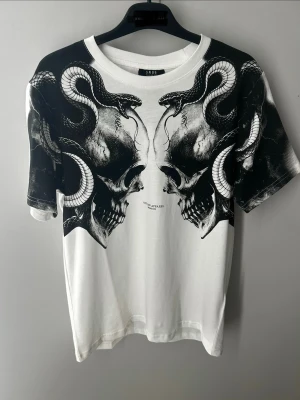 Vit t-shirt med dödskalle och ormar - Snygg vit t-shirt med stort svart tryck av två dödskallar och slingrande ormar på framsidan. T-shirten har rund halsringning och korta ärmar. Perfekt för dig som gillar edgy och unik stil. flera storlekar 
