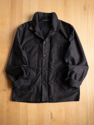 Le Laboureur Black Chore Jacket, size L - Stilren svart overshirt i kraftigt bomullstyg med klassisk krage och tre stora utanpåliggande fickor från kända franska varumärket.. Knäpps med fyra knappar framtill och har raka ärmar. Perfekt lager-på-lager-plagg för en avslappnad look.