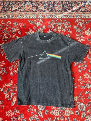 Svart Pink Floyd t-shirt med tryck - Cool svart t-shirt med ikoniskt Pink Floyd-tryck på bröstet. T-shirten har korta ärmar och en tvättad vintage-look. Materialet känns mjukt och skönt, troligtvis bomull. Perfekt för dig som gillar bandtröjor och vill sticka ut.