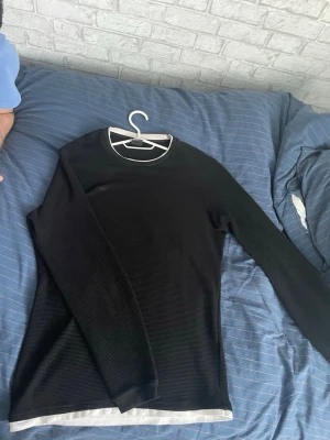 Svart långärmad tröja från FSBN - Svart långärmad tröja från FSBN i slim fit-modell. Tröjan har rund halsringning och vita detaljer vid krage och nederkant. Materialet är ribbat och ger en clean och stilren look. Perfekt för dig som gillar enkel och snygg stil.