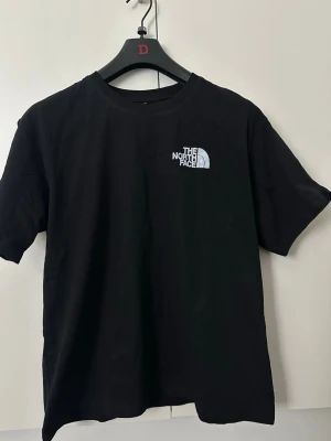 Svart t-shirt från The North Face - Klassisk svart t-shirt från The North Face med vit logga tryckt på bröstet. T-shirten har rund halsringning och korta ärmar. Tillverkad i mjuk bomull för en skön och avslappnad stil.
