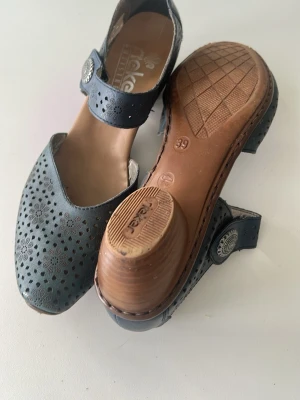 Mörkblå sandaler med låg klack - Mörkblå sandaler från Rieker i läder med dekorativt hålmönster och rundad tå. Skon har en bred rem med silverfärgad knappdetalj och låg klack i trälook. Yttersulan är i gummi för extra grepp och komfort. Perfekt för dig som gillar stilrena detaljer.