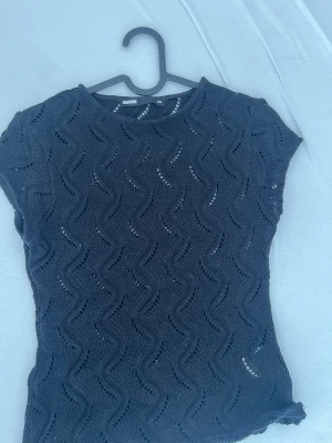 Svart virkad topp med kort ärm - Snygg svart virkad topp med vågigt hålmönster och korta ärmar. Toppen har rund halsringning och är lätt transparent tack vare det virkade materialet. Perfekt att styla med jeans eller kjol för en trendig look.