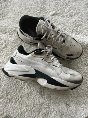 Vita chunky sneakers från Puma - Säljer ett par vita chunky sneakers från Puma med svarta detaljer. Skorna har en kraftig sula, meshpartier för extra ventilation och klassisk snörning. Perfekta för dig som gillar en sportig och trendig look. Lite nedtryckta i hälarna men inget jag känner när jag går