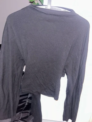 Svart långärmad croppad topp - Säljer en svart långärmad croppad topp med knyt baktill. Aldrig använd. Toppen har enkel design och är tillverkad i mjukt material, perfekt att styla med höga jeans eller kjol. Passar dig som gillar minimalistisk och trendig look.