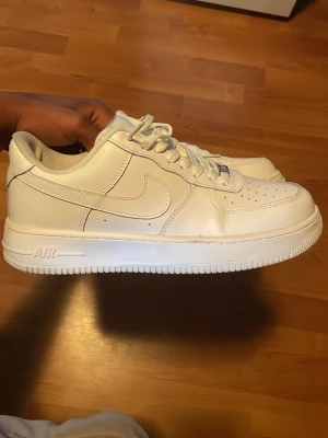 Vita Nike Air Force 1 sneakers - Klassiska vita Nike Air Force 1 sneakers i lågt utförande. Skorna är tillverkade i slätt läder med perforeringar på tån för ventilation och har den ikoniska swoosh-loggan på sidan. Tjock vit sula med AIR-detalj och vita skosnören.