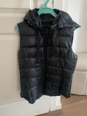 Svart dunväst med huva - Snygg svart dunväst med huva och dragkedja framtill. Lite glansigare i materialet. Passar till det mesta.