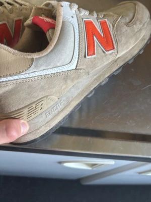 Beige New Balance sneakers med orange N - Säljer ett par beigea New Balance sneakers med orange N-logga på sidan. Skorna har ovandel i mocka och mesh, vita detaljer och grå sula. Klassisk retrostil med ENCAP-dämpning och lågt skaft. Perfekta för dig som gillar streetwear och snygga detaljer.