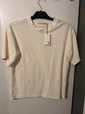  vit t-shirt från 157 - Säljer en ribbad vit t-shirt från 157 med rund halsringning och korta ärmar. T-shirten har en mjuk struktur och är perfekt för en clean och enkel stil. Materialet känns som bomull och ger en skön känsla mot huden. I storleken 160 cm. (Aldrig använt)