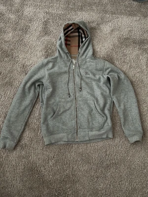 Burberry hoodie - Snygg grå hoodie med dragkedja framtill och två stora fickor. Insidan av huvan och öppningen har ett klassiskt rutigt mönster i beige, svart och rött. Tillverkad i mjukt sweatshirtmaterial och har justerbara snören i huvan. Perfekt för en chill och avslappnad stil.