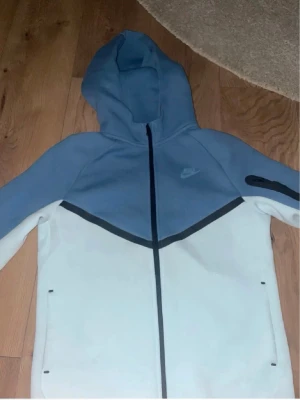 Nike blå och vit hoodie med dragkedja - Nike hoodie i ljusblått och vitt med hel dragkedja framtill. Tröjan har huva, svarta detaljer och två fickor med dragkedja. Nike-logga på bröstet och en svart detalj på ena ärmen. Perfekt för en sportig och avslappnad stil.