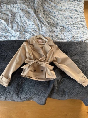 Kort trenchcoat  - Säljer denna trenchcoat från Gina 14+. Slutsåld och extremt eftertraktad. Storlek M men är kort i modellen så passar bättre som S/XS. Använd 1 gång❤️❤️❤️