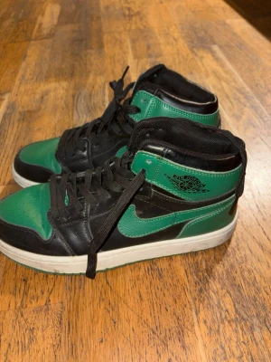 Äkta Svart och grön Air Jordan 1 High - Snygga Air Jordan 1 High sneakers i svart och grönt läder med klassisk vit sula. Skorna har svarta skosnören, gröna detaljer på tå, sidor och häl samt ikoniska Air Jordan-loggan på sidan. Perfekta för dig som vill sticka ut med stilrena och sportiga sneakers.