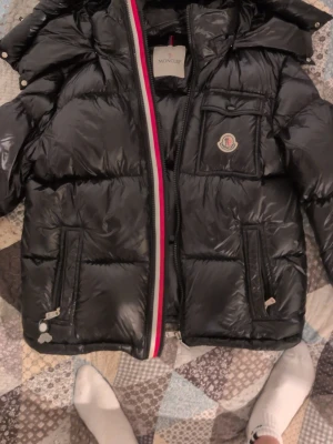 Svart dunjacka från Moncler - Snygg svart dunjacka från Moncler med glansig finish, ikonisk logga på bröstet och randig detalj längs dragkedjan. Jackan har flera fickor med dragkedja och en innerficka med serietidningsmönster. Perfekt för kalla vinterdagar.