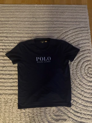 Mörkvblå t-shirt från Polo Ralph Lauren - Snygg mörkblå t-shirt från Polo Ralph Lauren med klassisk logga tryckt i vitt på bröstet. T-shirten har rund halsringning och korta ärmar. Perfekt till jeans eller shorts för en clean och stilren look.