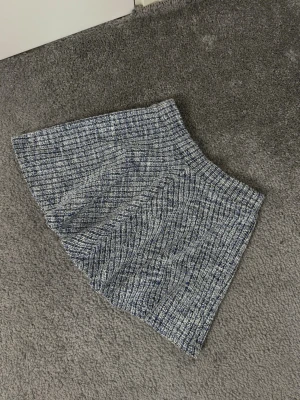 Blått tweed set med väst och shorts - Säljer ett snyggt set i blått och vitt tweedtyg, bestående av en ärmlös väst med guldknappar och två fickor framtill samt matchande shorts med hög midja. Perfekt för en trendig och chic look.