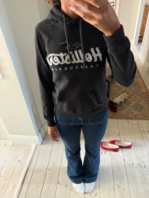 Svart Hollister hoodie - Svart hoodie från Hollister med stor broderad logga och texten 'California' på bröstet. Klassisk känguruficka framtill, ribbade muddar och dragsko i huvan. Mjuk bomullsblandning som är skön att ha på sig. Perfekt för chill dagar eller när du vill ha en avslappnad look.