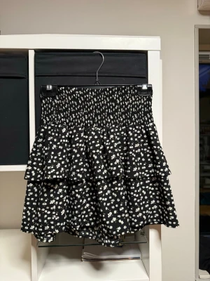 Svart volangkjol med vita blommor - Kort svart volangkjol med vita småblommiga mönster och bred smock i midjan. Har använts få gånger. Storlek 158/164 men sitter som en vanlig kjol på mig som har xs-s. Går också att ha som tubtopp! 