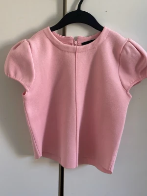 Ljusrosa topp med puffärm - Supersöt ljusrosa topp med puffärmar och rund hals. Toppen har en rak passform och dragkedja i ryggen för enkel påklädning. Perfekt för dig som gillar en feminin och stilren look.