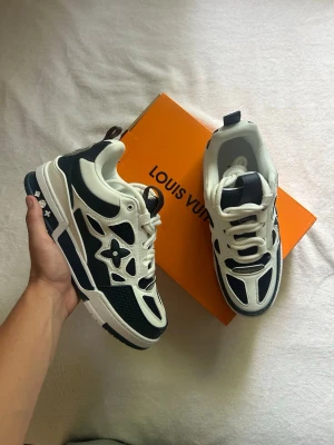 Louis Vuitton vita och blåa sneakers - Snygga Louis Vuitton sneakers i vitt och blåty med ikoniska LV-detaljer och blommor på sidan. Skorna har en chunky sula med tydlig logga undertill och meshpaneler för extra ventilation. Perfekt för dig som vill sticka ut med exklusiva detaljer och modern design.