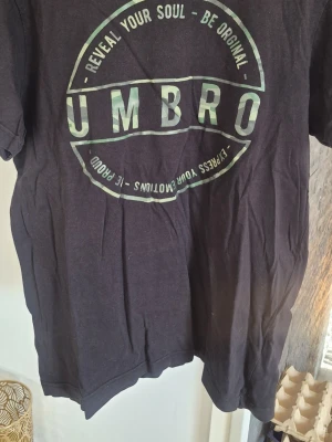 Svart t-shirt från Umbro - Säljer en svart t-shirt från Umbro med stort tryck framtill i grönt och vitt. T-shirten har klassisk passform och rund hals. Perfekt för dig som gillar sportiga och avslappnade plagg.