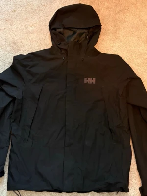 Svart skidjacka från Helly Hansen - Svart skidjacka från Helly Hansen med huva och dragkedja framtill. Jackan har två stora fickor på framsidan och justerbar nederkant. Tillverkad i vattenavvisande material, perfekt för regniga och snöiga dagar. Diskret HH-logga på bröstet.