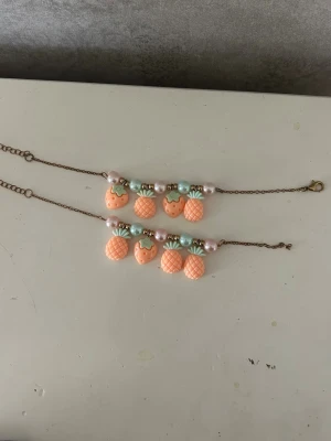 Söta armband med fruktberlocker - Två armband med kedja i bronsfärg och berlocker formade som jordgubbar och ananaser i ljusrosa och orange. Armbanden har även pärlor i pastellfärger och små gulddetaljer. Perfekt accessoar för dig som gillar lekfulla smycken.