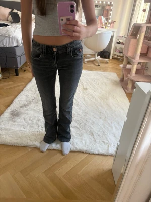Mörkgrå bootcut jeans med brodyr - Säljer ett par mörkgrå bootcut jeans med snygga broderade detaljer på bakfickorna. Jeansen har låg midja och klassisk femficksdesign. Perfekta för dig som gillar en avslappnad men ändå trendig stil.
