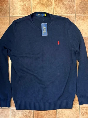 Polo Ralph Lauren stickad tröja – L - Helt ny Polo Ralph Lauren stickad tröja i marinblå/navy. Aldrig använd och kommer med originaltags kvar. Klassisk modell med den ikoniska röda Polo Ralph Lauren-loggan på bröstet. Stilren och lättmatchad