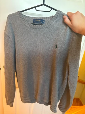 Grå stickad tröja Polo Ralph Lauren - Klassisk grå stickad tröja från Polo Ralph Lauren med rund halsringning och diskret broderad logga på bröstet. Tröjan har långa ärmar och ribbade muddar vid ärmslut och nederkant. Perfekt basplagg till garderoben.