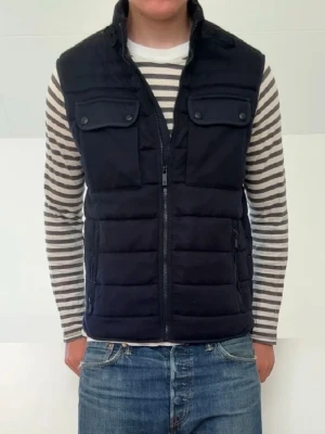 Massimo duktig väst - Snygg svart dunväst med ståkrage och dragkedja framtill. Västen har två bröstfickor med lock och knappar samt två sidofickor. Quiltad design som ger en modern och sportig look. Perfekt att slänga över en tröja för extra värme.