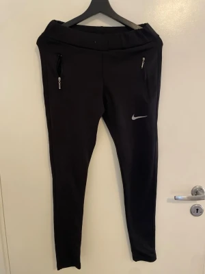 Svarta träningsbyxor från Nike - Säljer ett par svarta träningsbyxor från Nike med dragkedjor på fickorna och den klassiska Nike-loggan på benet. Byxorna har smal passform och är perfekta för löpning eller gym. Tillverkade i ett stretchigt material för bästa komfort.