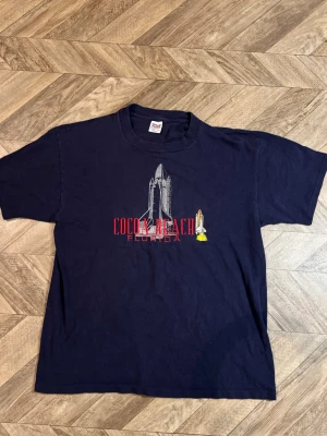 Mörkblå Cocoa Beach Florida t-shirt - Mörkblå t-shirt med tryck av en rymdraket och texten 'Cocoa Beach Florida' på bröstet. Klassisk rund hals och korta ärmar. Perfekt för dig som gillar rymd eller vill ha en cool souvenirtröja.