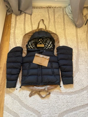 Burberry jacka &väst  - Burberry Dunjacka med avtagbara ärmar  Exklusiv Burberry-dunjacka med klassisk Burberry-rutig huva och avtagbara ärmar, vilket gör att den även kan användas som väst.  Anmärkningar:  Dragkedjan på en ficka har byts bytas • Lagning finns på nedersta panelerna på båda ärmarna  I övrigt är jackan i mycket fint skick med bra dunfyllning och passform. Nypris ligger på ett mycket högt belopp, vilket gör detta till ett prisvärt alternativ för dig som vill bära Burberry. 🔥