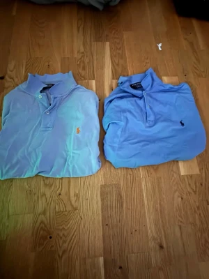 Blå pikétröjor från Polo Ralph Lauren - Två klassiska blå pikétröjor från Polo Ralph Lauren med broderad logga på bröstet, en med orange och en med marinblå logga. Tröjorna har krage, knappar och är tillverkade i bomull. Perfekt för en stilren och avslappnad look. Har använt båda i flera år.