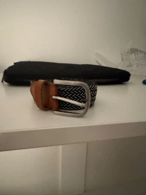 Flätat bälte - Snyggt svartvitt flätat bälte med bruna läderdetaljer och silverfärgat spänne. 110cm