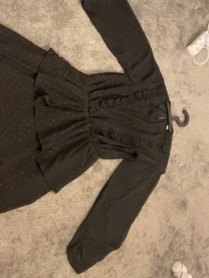 Svart prickig volangklänning - Snygg svart klänning med små prickar och volangdetaljer vid midjan och längs urringningen. Klänningen har lång ärm och markerad midja som ger en feminin siluett. Perfekt för dig som gillar en klassisk men lekfull stil.