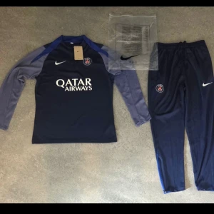 PSG träningsset från Nike, mörkblå - Snyggt träningsset från Nike för PSG med långärmad tröja och byxor i mörkblått. Tröjan har halv dragkedja, PSG-logga och Qatar Airways-tryck på bröstet. Byxorna har PSG-märke och Nike-logga. Perfekt för fotbollsträning eller chill.