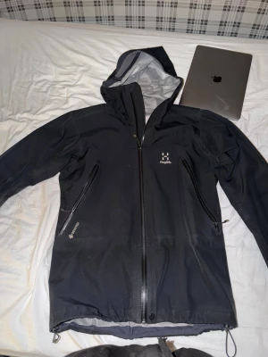 Svart regnjacka från Haglöfs, Gortex - Snygg svart regnjacka från Haglöfs med hel dragkedja framtill, två stora snedställda fickor med dragkedja och justerbar huva. Jackan har minimalistisk design och är tillverkad i ett vattentätt material, perfekt för regniga dagar. Passar även M.