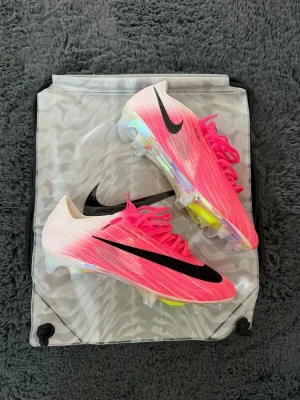Nike Mercurial rosa fotbollsskor - Säljer ett par Nike Mercurial fotbollsskor i en grymt snygg rosa färg med svart swoosh. Skorna har en gradient från vitt till rosa, genomskinlig yttersula med neon-detaljer och snörning. Tillverkade i lätt syntetmaterial för maxad speed på planen.