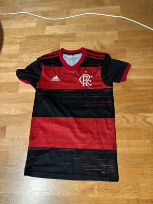 Svart och röd Flamengo matchtröja - Säljer en svart och röd randig Flamengo matchtröja från Adidas med klubbmärke och Adidas-logga på bröstet. Tröjan har V-ringning, korta ärmar och detaljer i vitt samt tryck på ryggen. Perfekt för fotbollsträning eller att bära som supporter.