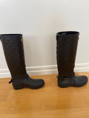 Svarta quiltade stövlar Barbour - Snygga svarta stövlar från Barbour med quiltat mönster på skaftet och dekorativt spänne vid ankeln. Tillverkade i gummi, perfekta för regniga dagar. Klassisk hög modell med Barbour International-logga upptill.