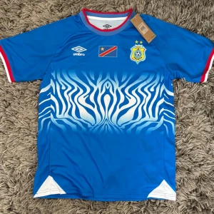Blå DR Kongo matchtröja från Umbro - Snygg blå matchtröja från Umbro med DR Kongos flagga och fotbollsförbundets emblem på bröstet. Tröjan har ett unikt zebramönster i vitt och blått framtill, röda och vita detaljer vid ärmslut och krage samt sportig passform. Perfekt för fotboll eller streetwear.