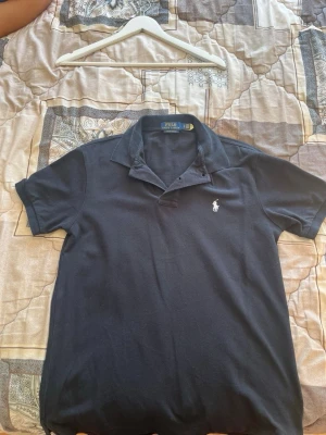 Mörkblå pikétröja från Polo Ralph Lauren - Klassisk mörkblå pikétröja från Polo Ralph Lauren med korta ärmar och vit broderad logga på bröstet. Tröjan har krage och knappar vid halsen. Perfekt till jeans eller shorts för en clean och stilren look.